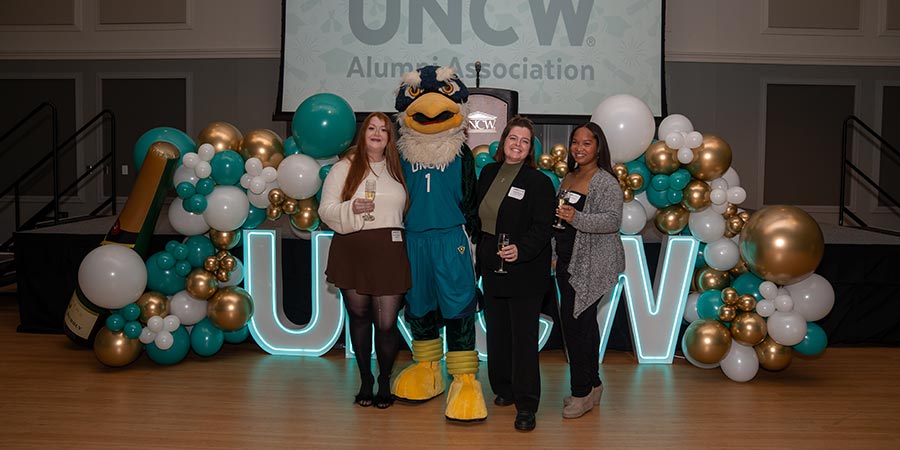 Three alumni pose with Sammy C. Hawk for a photo